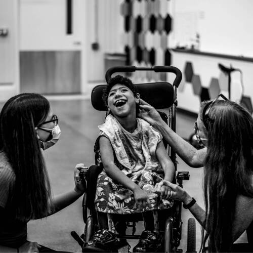 Pictured right, Summer Scholar Brianna Kleikamp, left, and Lisa Kenyon, professor of physical therapy, right, congratulate Gabby, center, for working hard during her powered mobility training using the IndieTrainer system at the Shape Corp. Innovation...